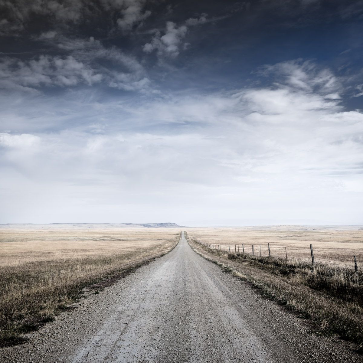 A gravel road stretches endlessly through vast prairie grasslands under dramatic cloudy skies.