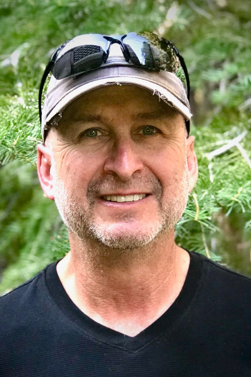 A person wearing sunglasses and a black baseball cap smiles while standing outdoors against a green foliage background.