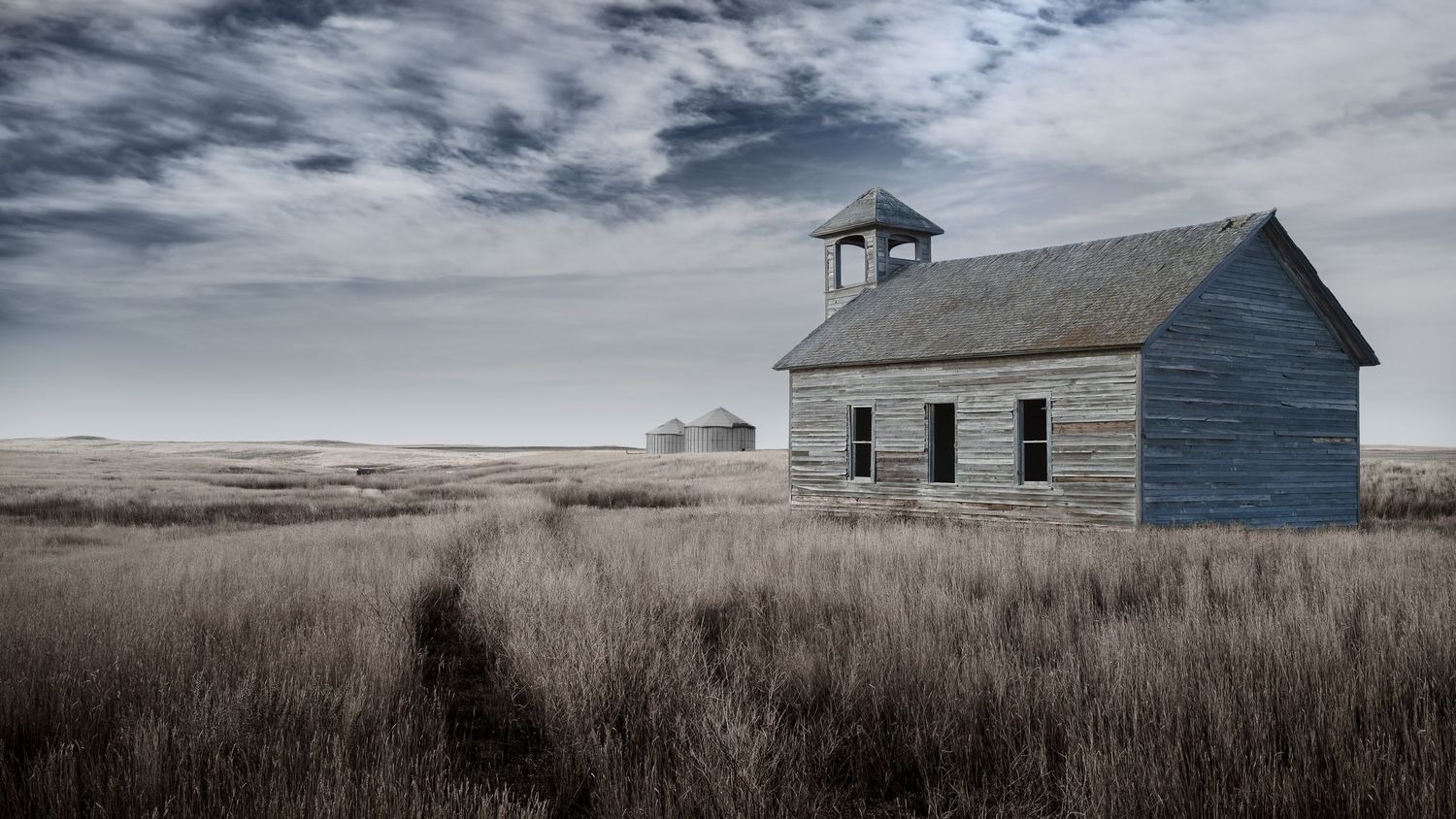 Abandoned wooden schoolhouse with bell tower stands in vast prairie grassland under dramatic cloudy sky.
