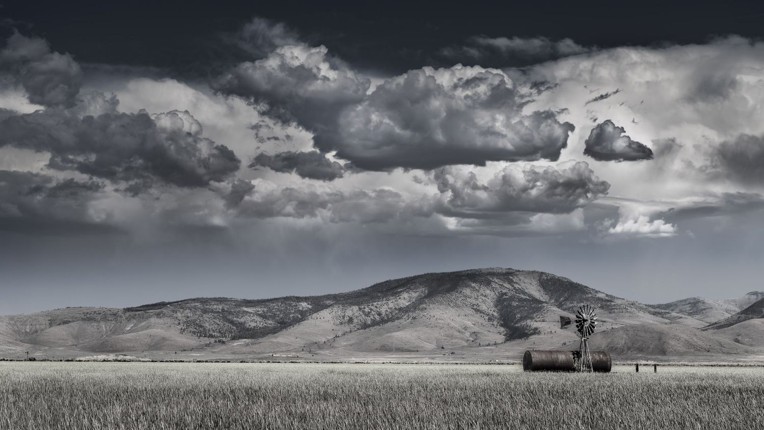 Black and white rural landscape with windmill, barn, rolling hills, and dramatic storm clouds over open grassland.
