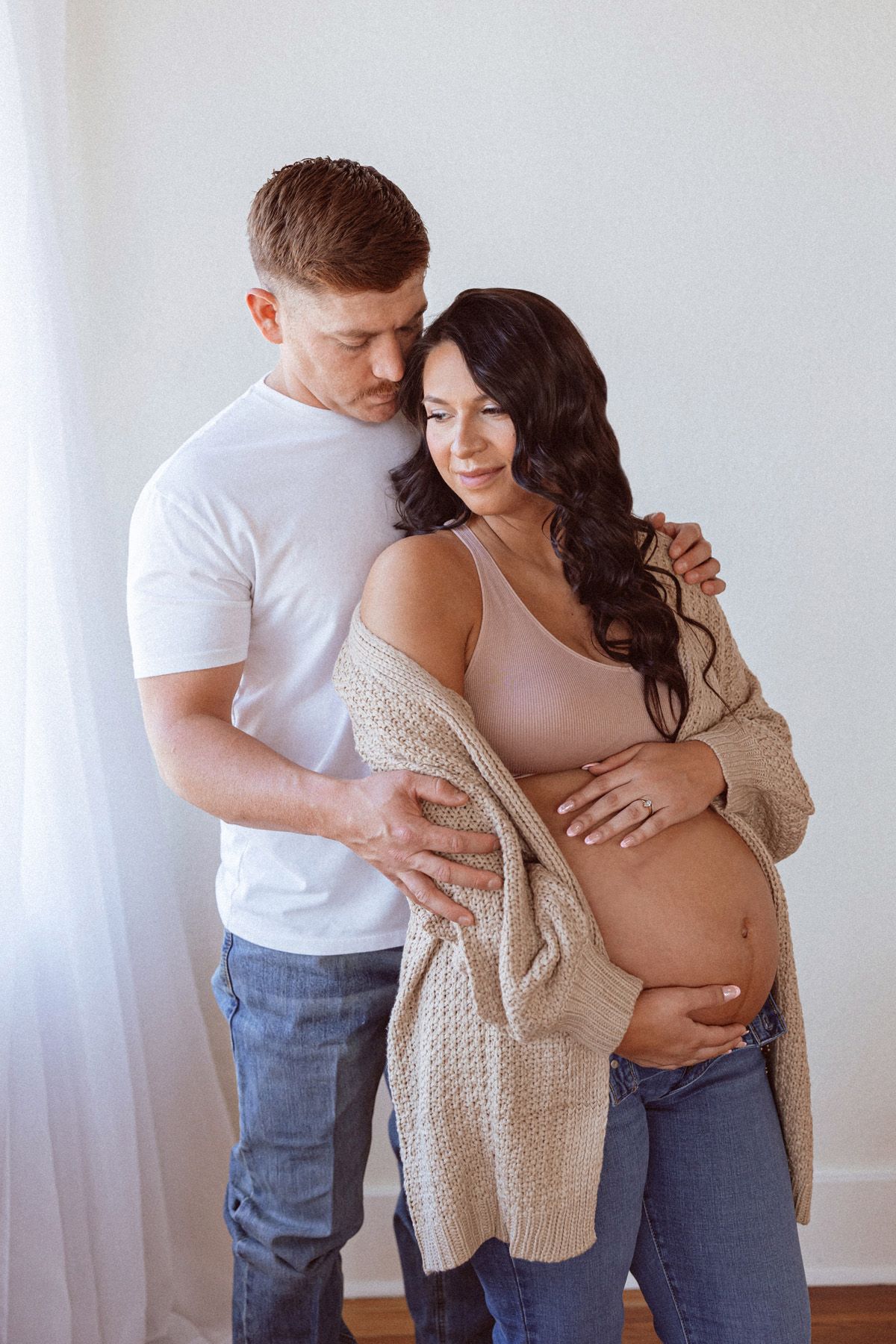 Intimate maternity photoshoot showing couple embracing in casual outfits against white wall.