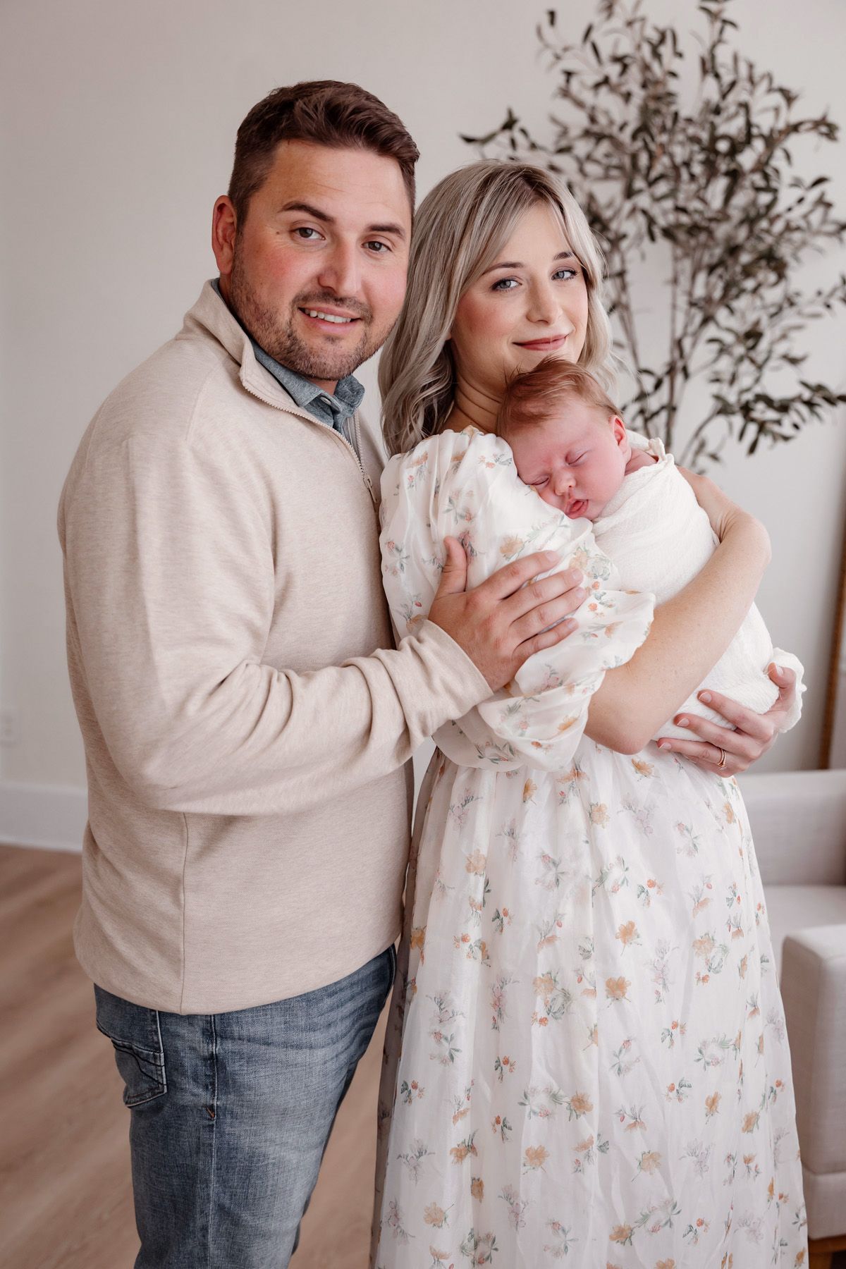 Sweet family moment with parents in casual clothes holding their newborn baby in a floral dress.