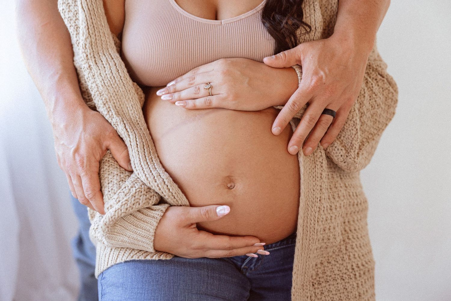 Close-up maternity photography showing gentle embrace and beige cardigan details.