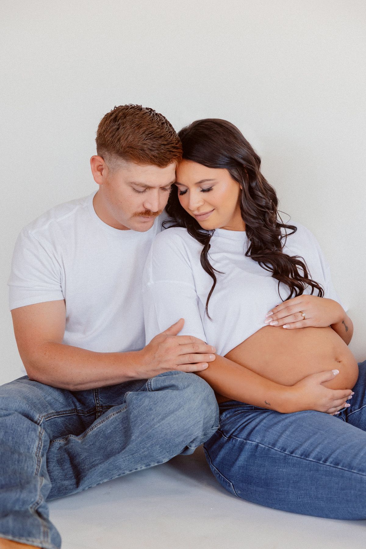 Intimate maternity photo of couple in white shirts and jeans sitting on floor.