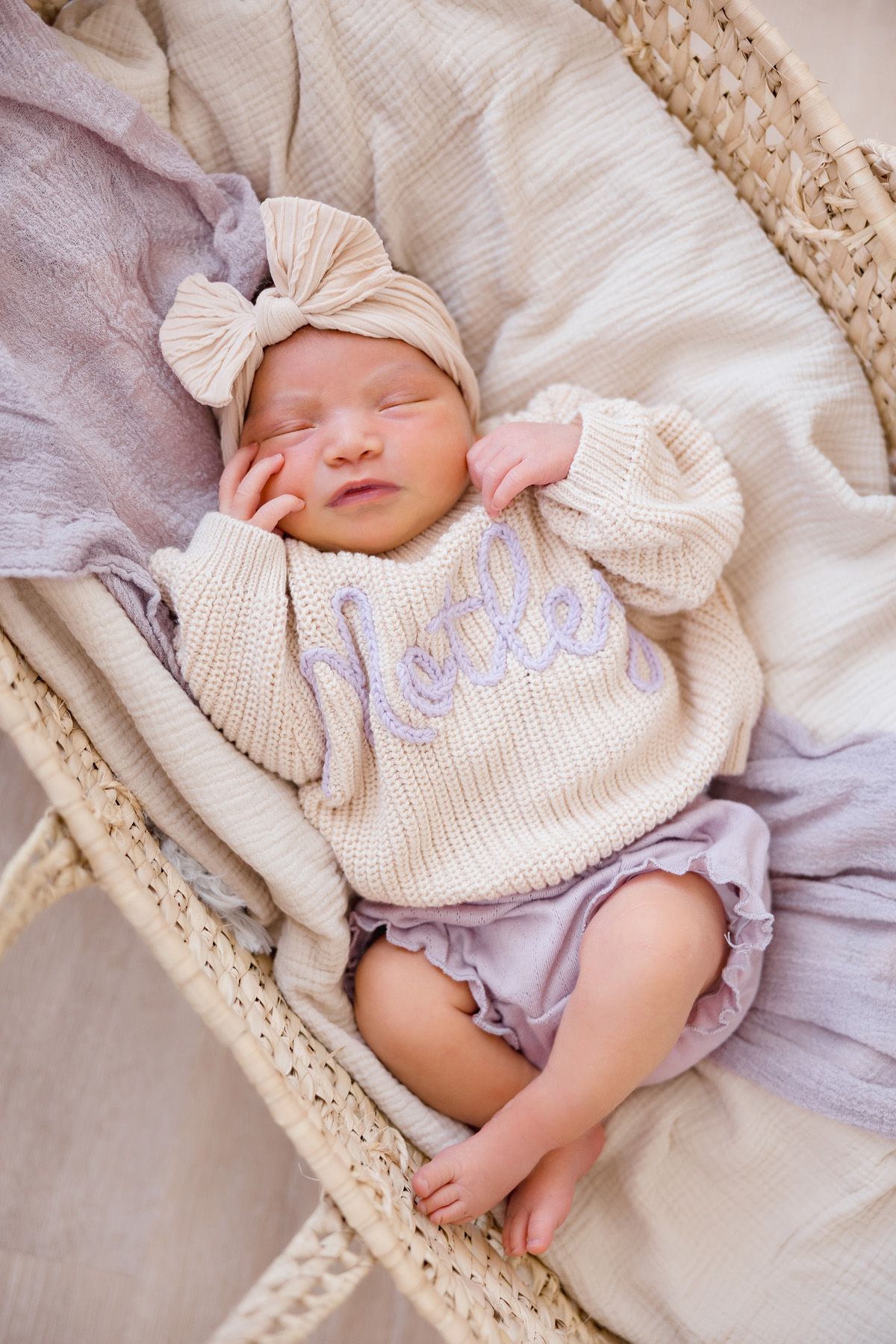 Sleeping newborn baby wearing cream knit sweater and bow rests peacefully on soft blankets.