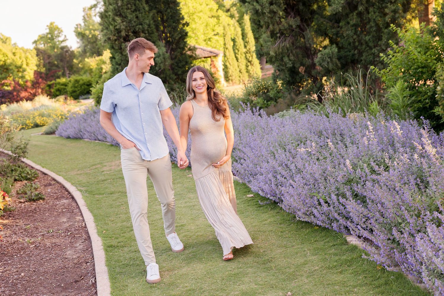 Couple walking through garden with lavender flowers during sunset maternity session.
