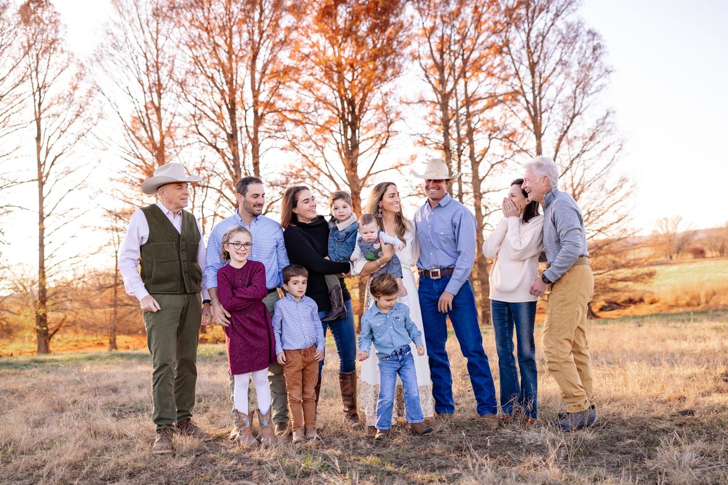 Extended family gathering poses for photos outdoors against a backdrop of bare trees at sunset.
