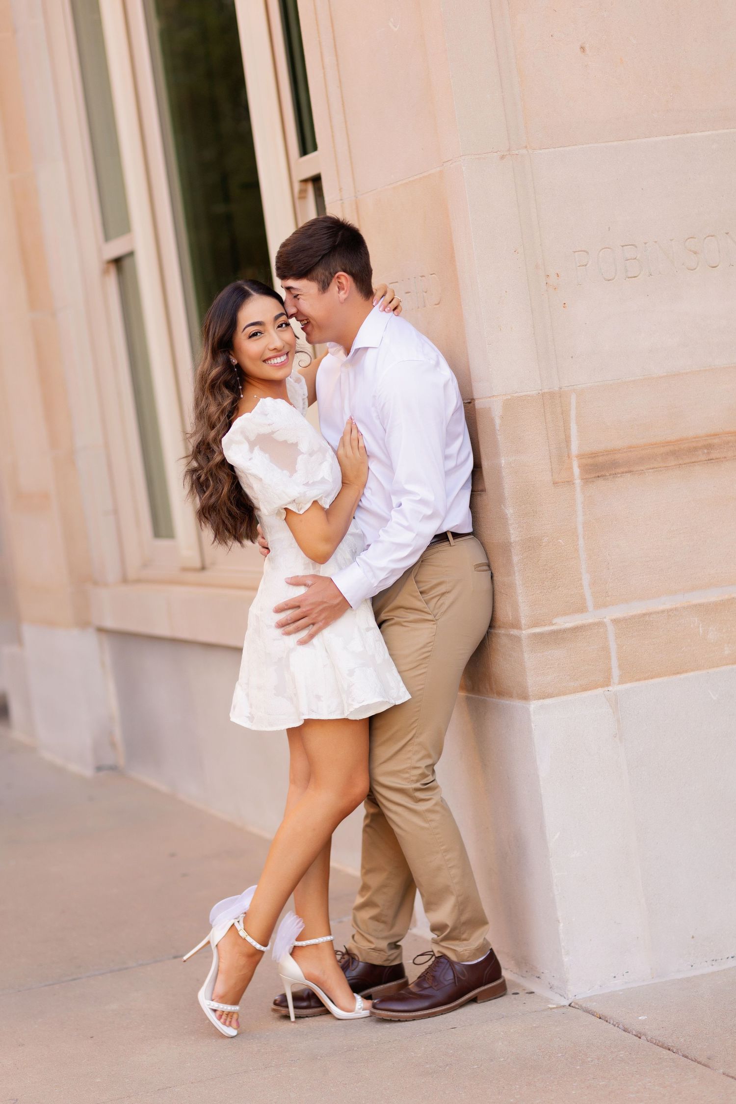 Romantic couple in white outfits share intimate moment against stone building wall.