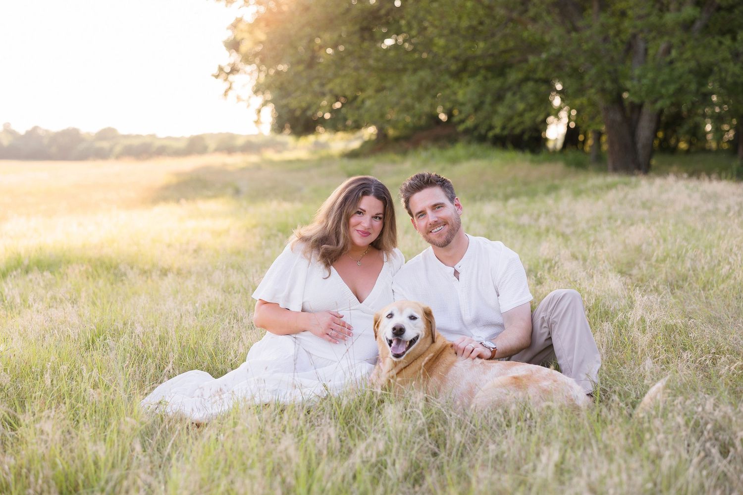 Family portrait in field at sunset with golden retriever dog sitting beside couple in white outfits.