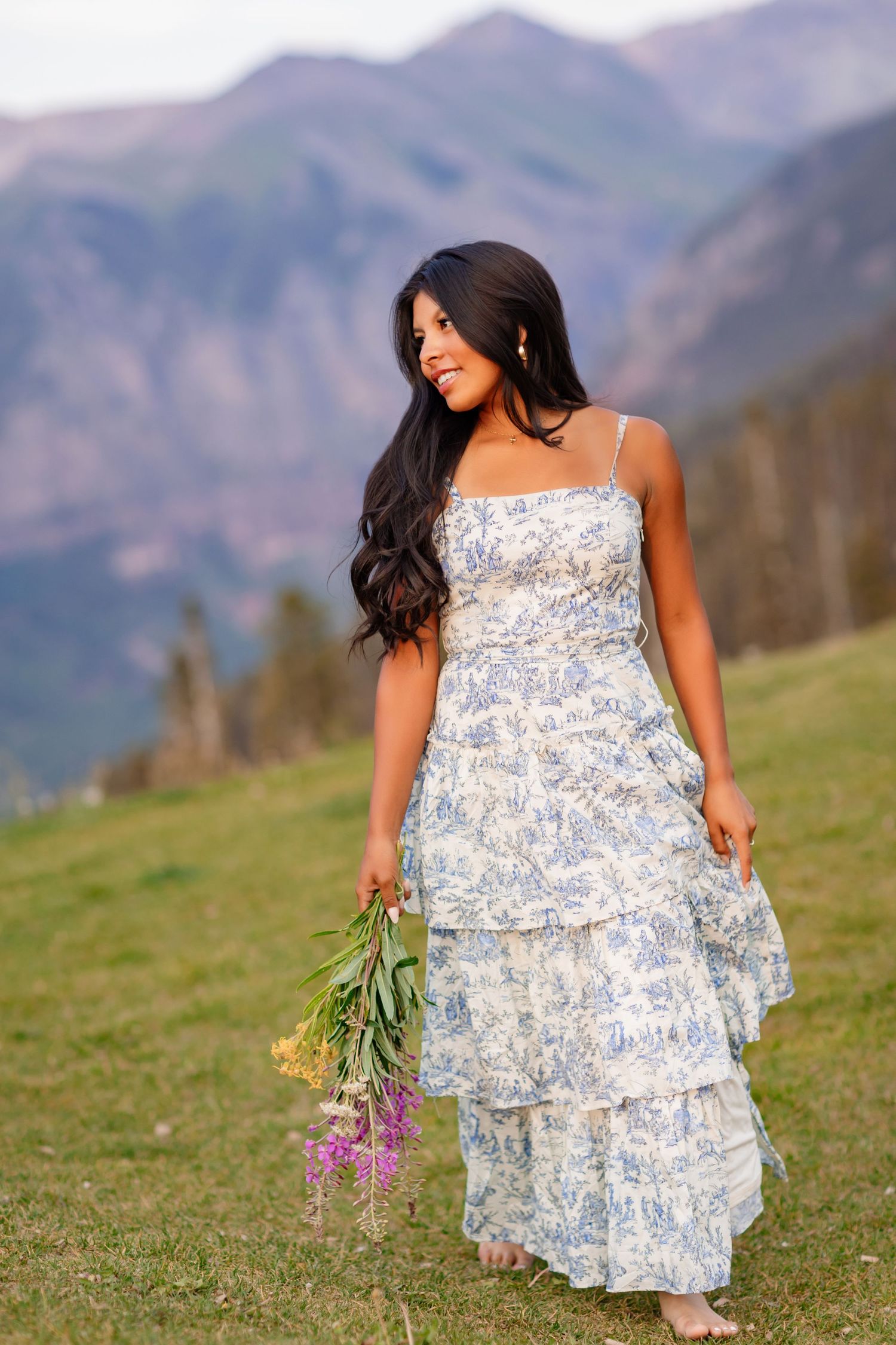 Person in flowing blue floral dress poses against mountain landscape with wildflowers.