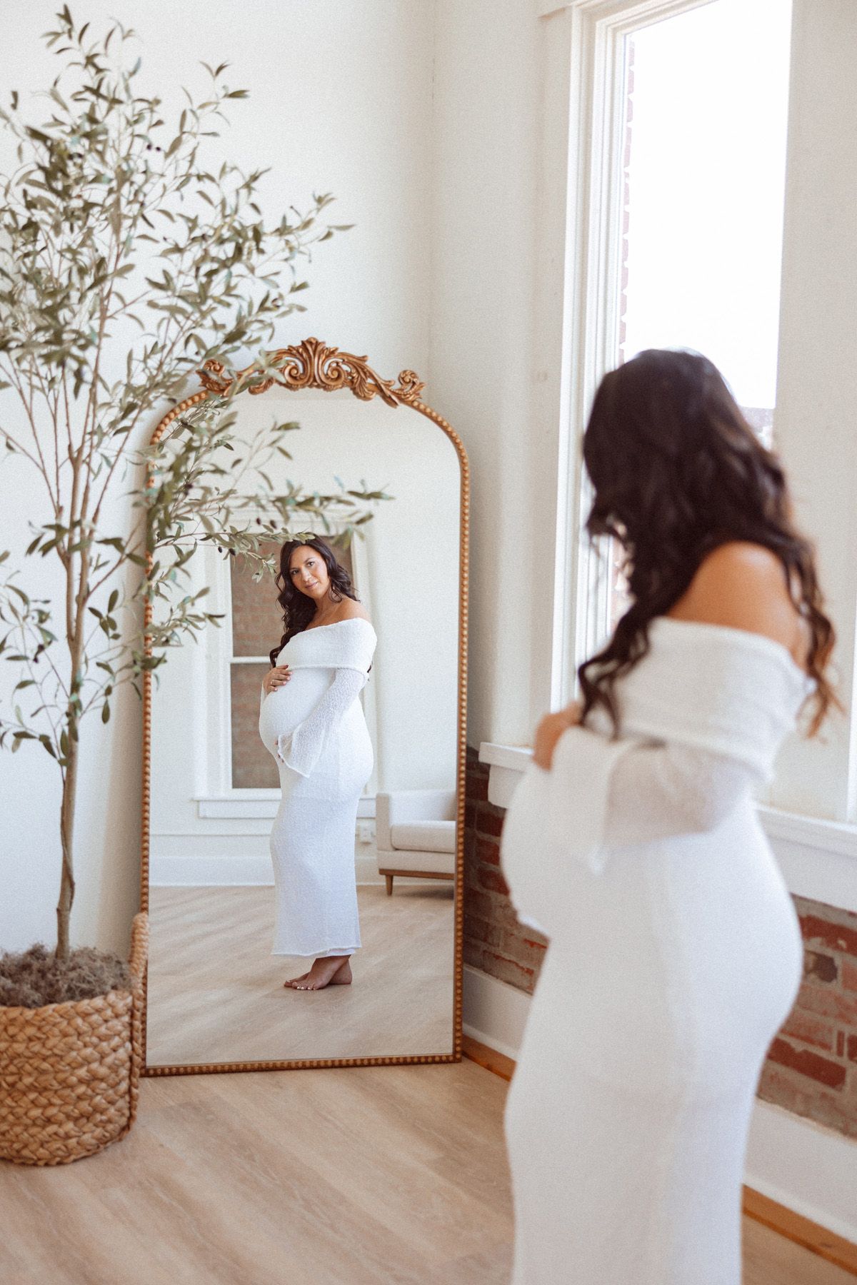 Maternity photos in white dress near window with decorative olive tree.