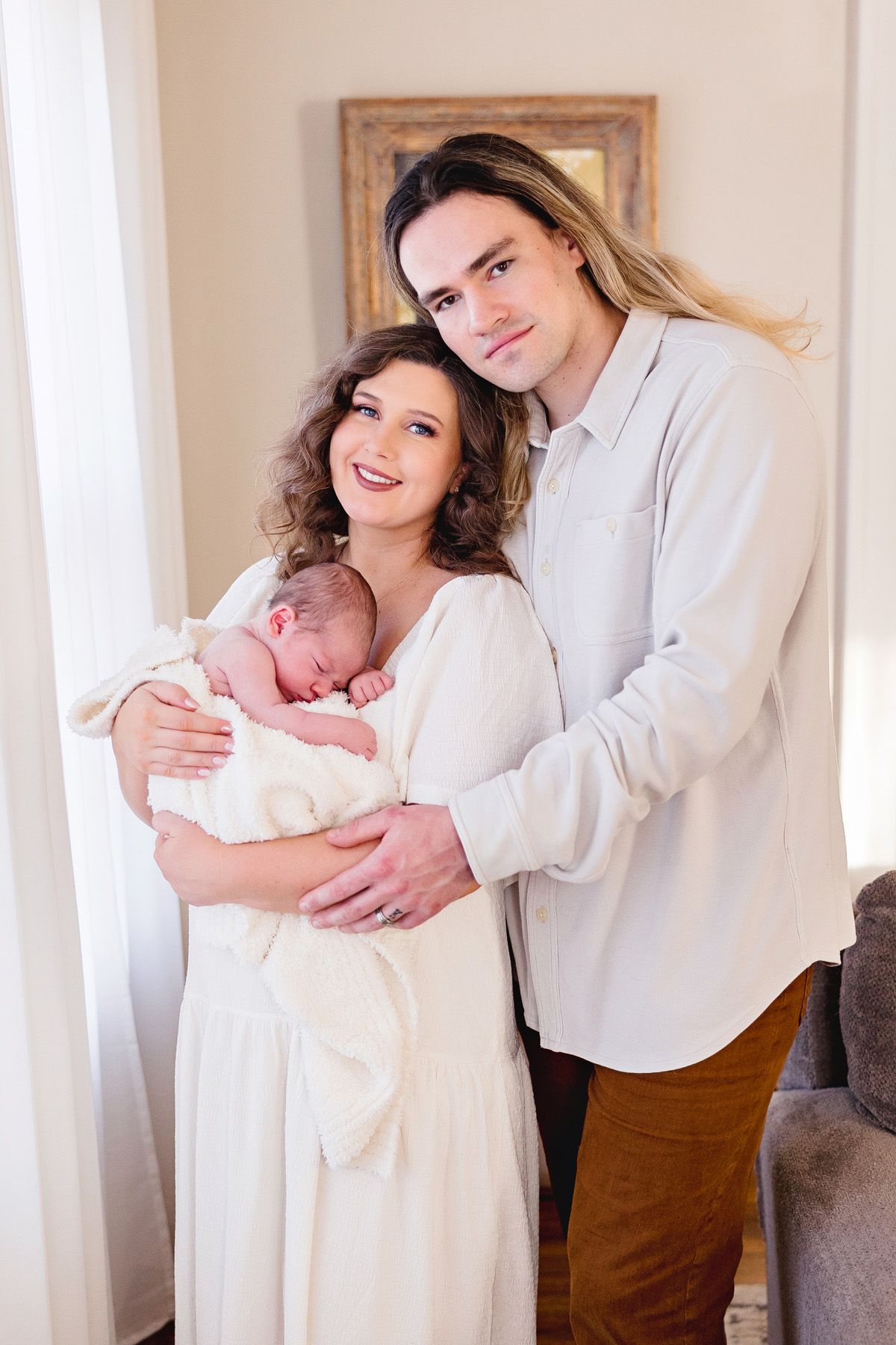 A couple in white outfits holds their newborn baby in a bright indoor setting with a framed picture in background.