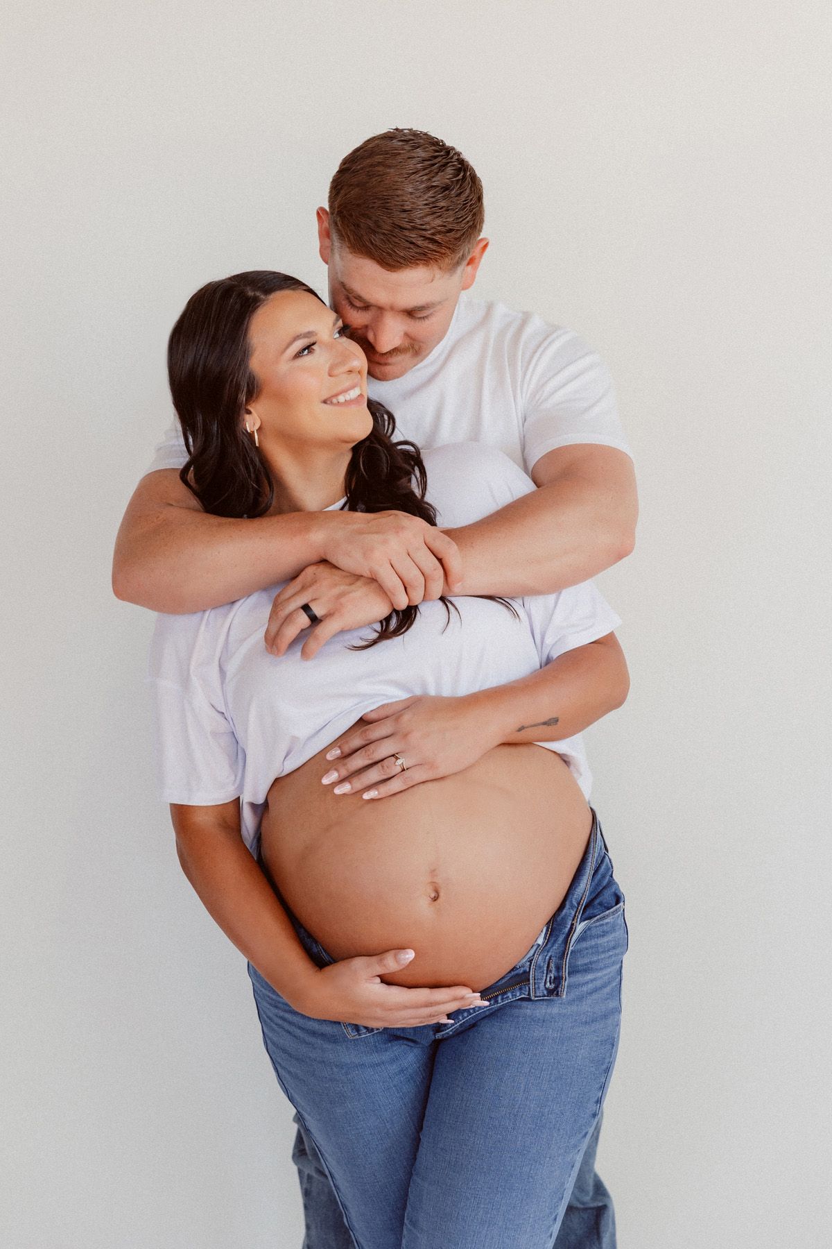 Intimate pregnancy portrait of couple embracing against white backdrop.