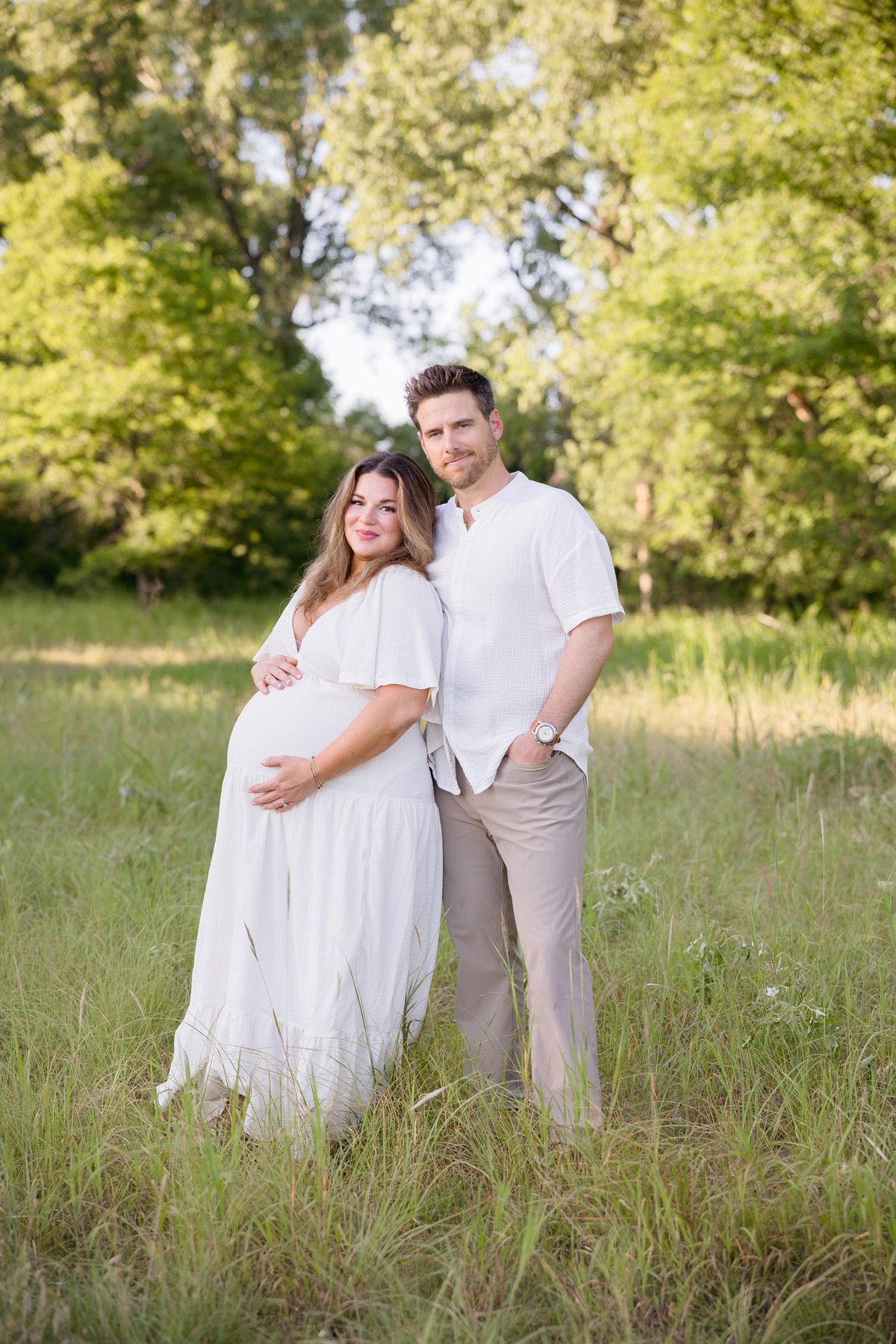 Maternity photo of a couple in white outfits standing together in a sunlit field.
