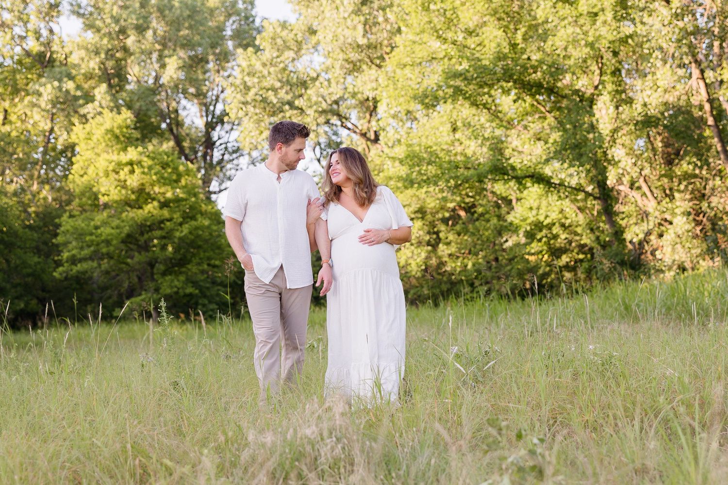Couple in white clothing walking together in a sunlit field with trees in background.