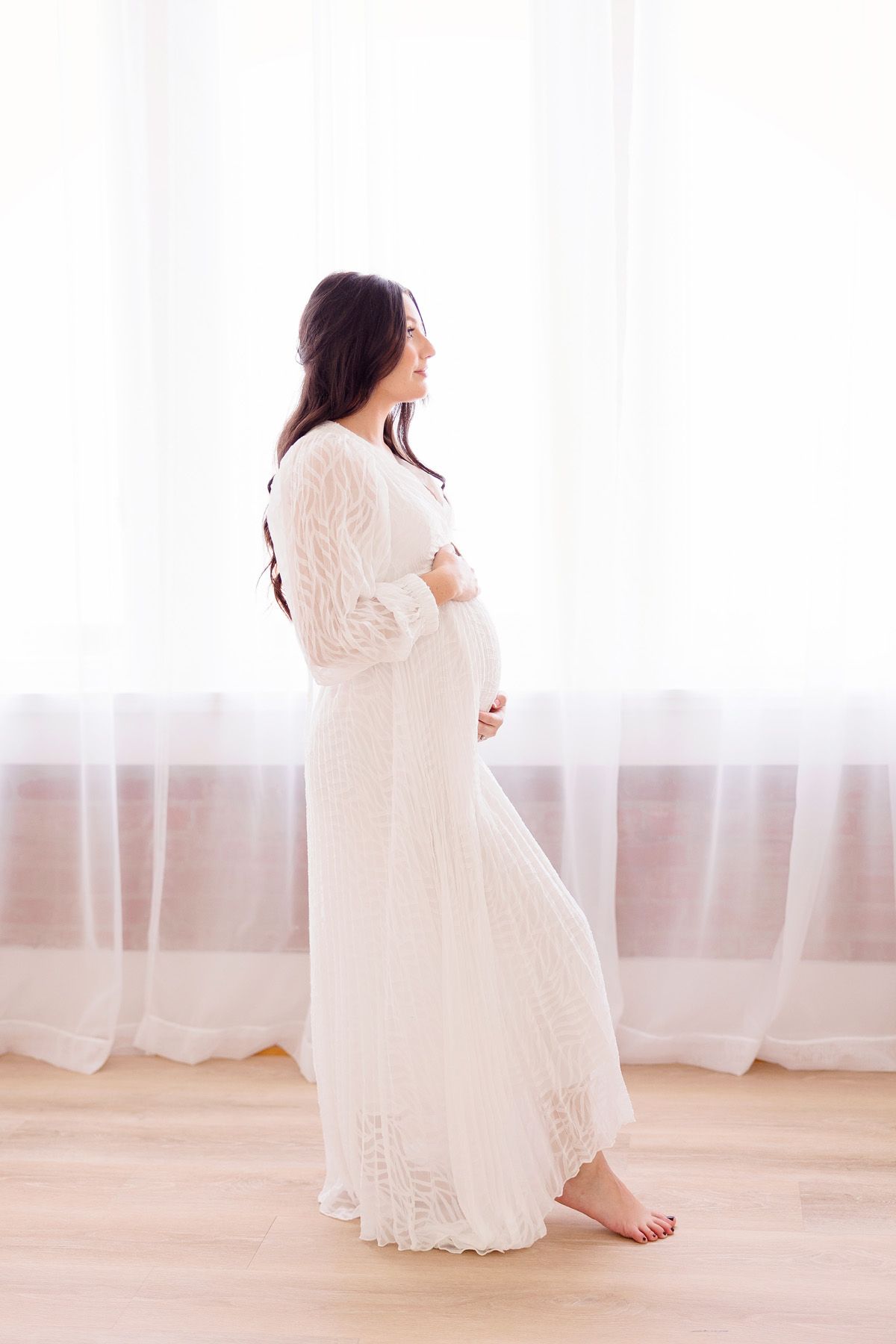 Figure in white lace dress poses gracefully against bright window backdrop oklahoma based studio photographer