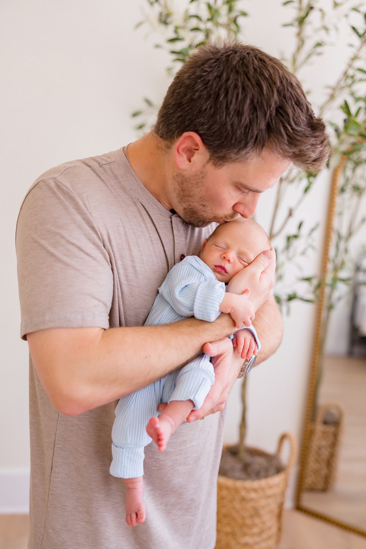 Parent tenderly kisses newborn baby wearing blue onesie while standing near indoor plant.