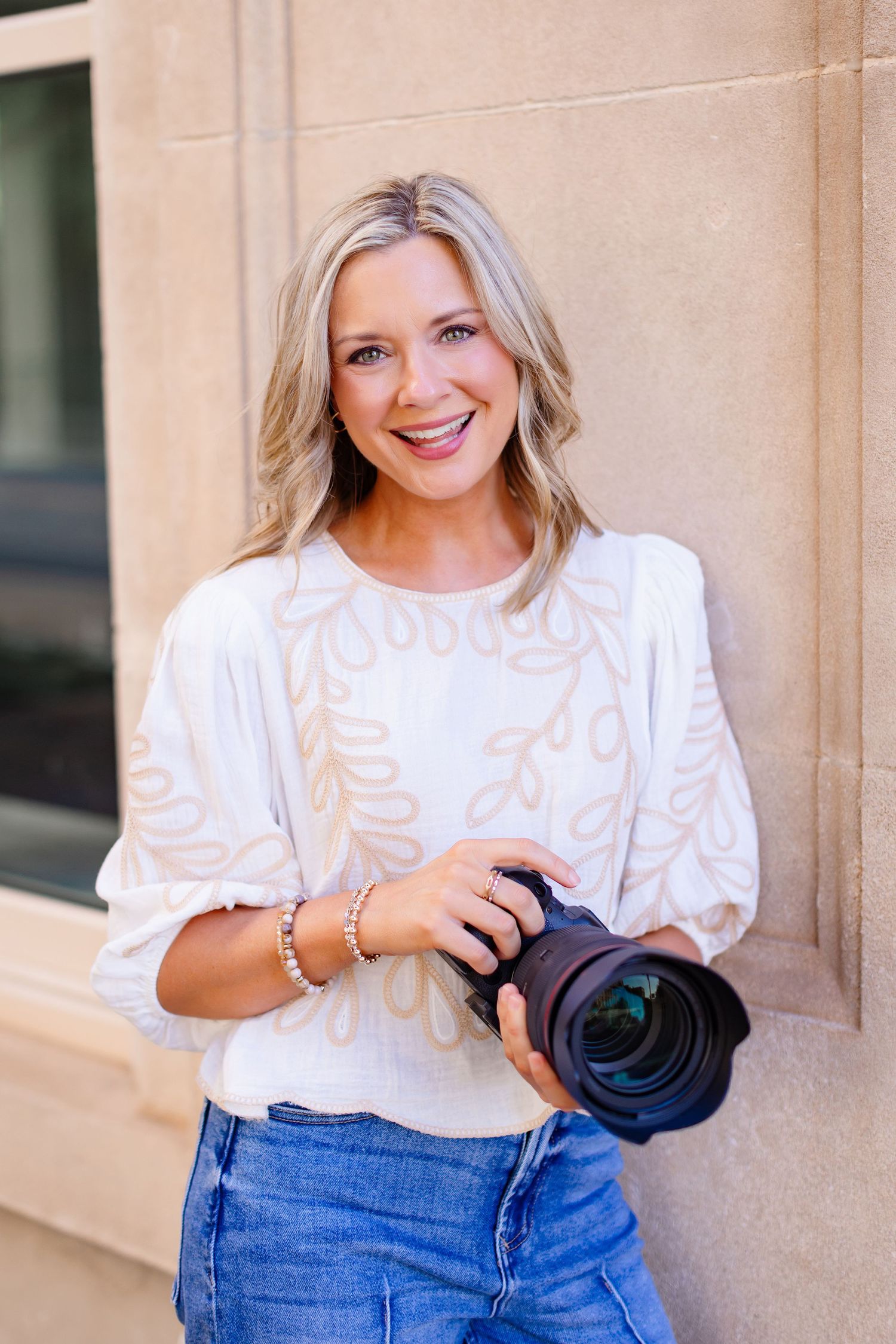 Professional photographer in white top and jeans poses with camera against stone wall.