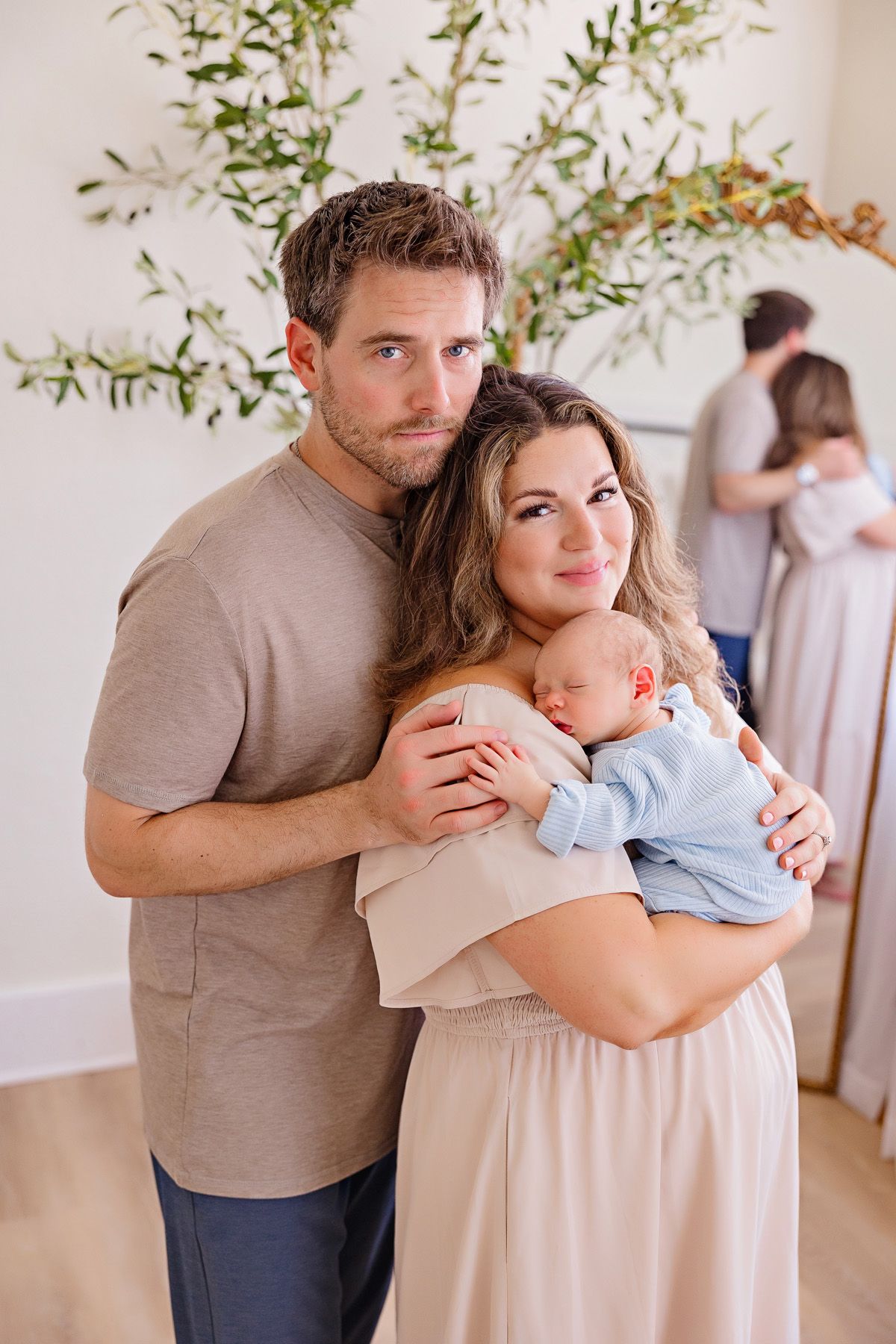 Indoor family portrait with newborn baby in blue outfit against white wall with plant decor.