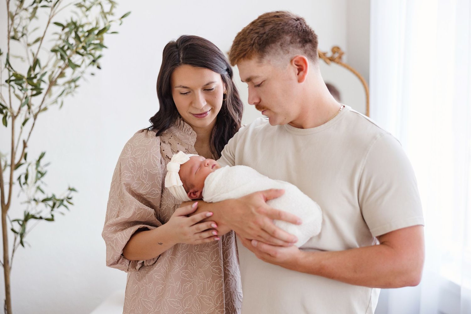 Couple in white shirts holding newborn baby while standing near green plants against white wall.