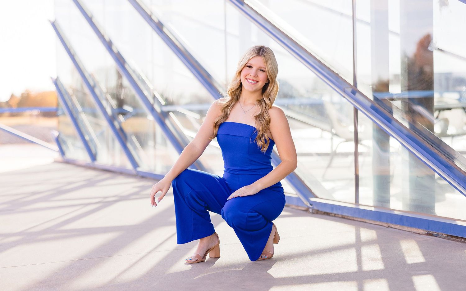 A dancer in a vibrant blue dress poses gracefully in a sunlit studio with glass windows and railings.
