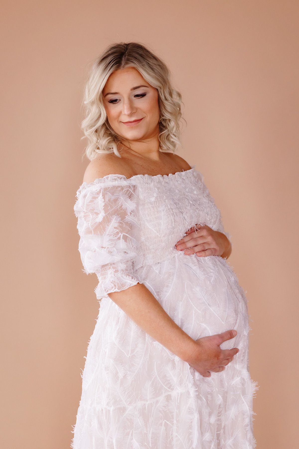 Maternity portrait session in elegant white lace dress against neutral backdrop.
