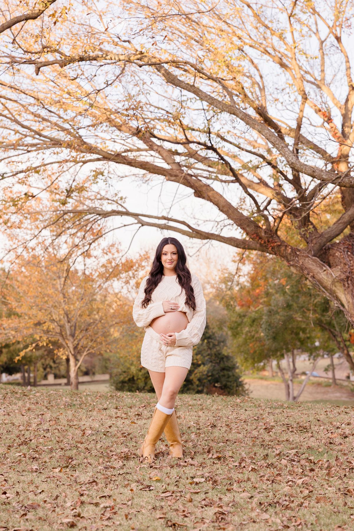 Maternity photos under autumn trees with fallen leaves creating natural backdrop.