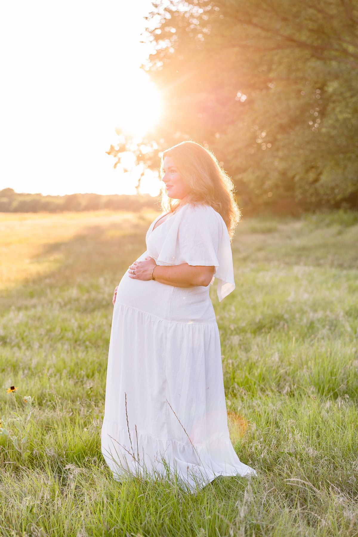 Maternity photo of a woman in a flowing white dress standing in a sunlit grassy field at golden hour.