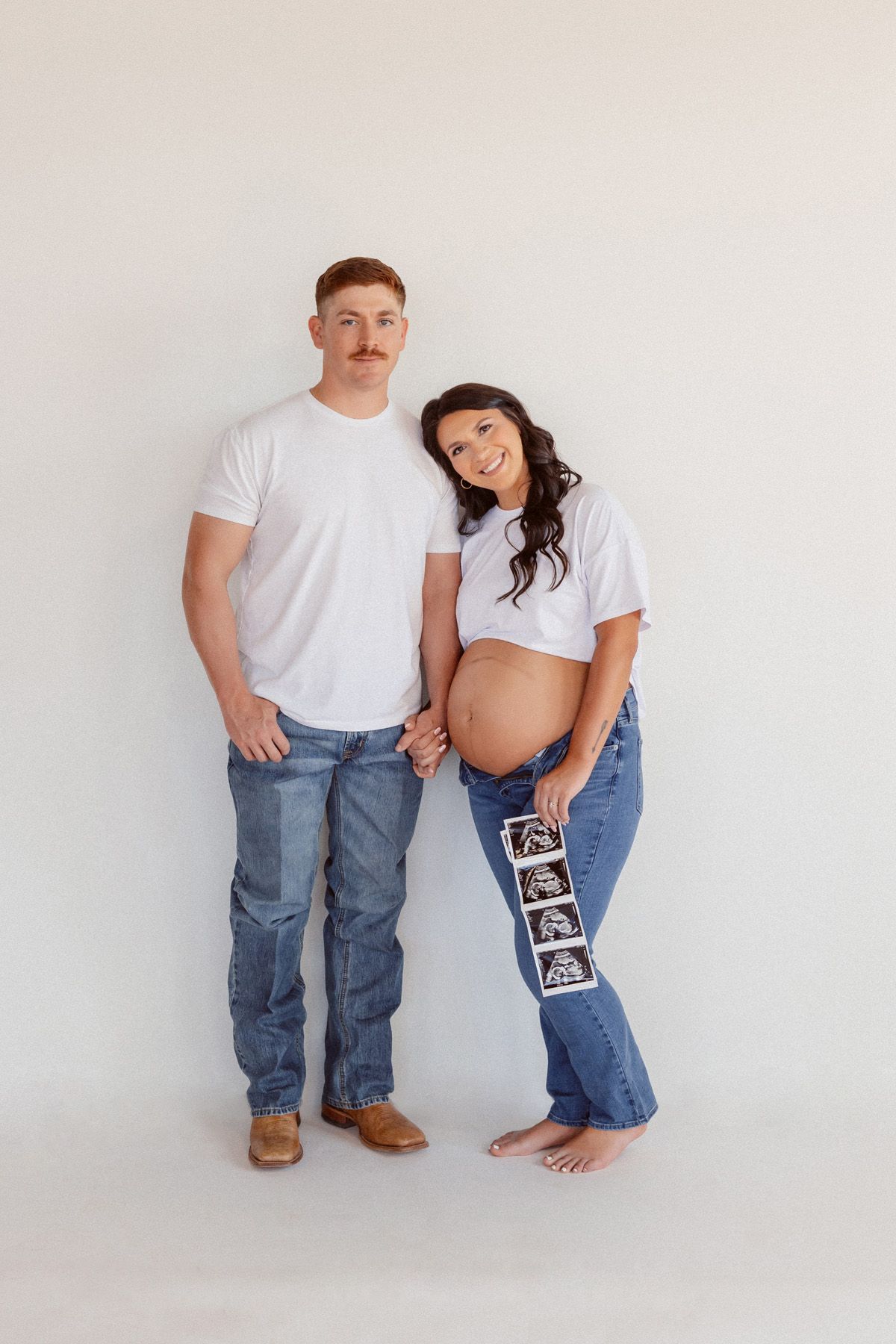 Barefoot couple in matching outfits pose with ultrasound photos against white background.