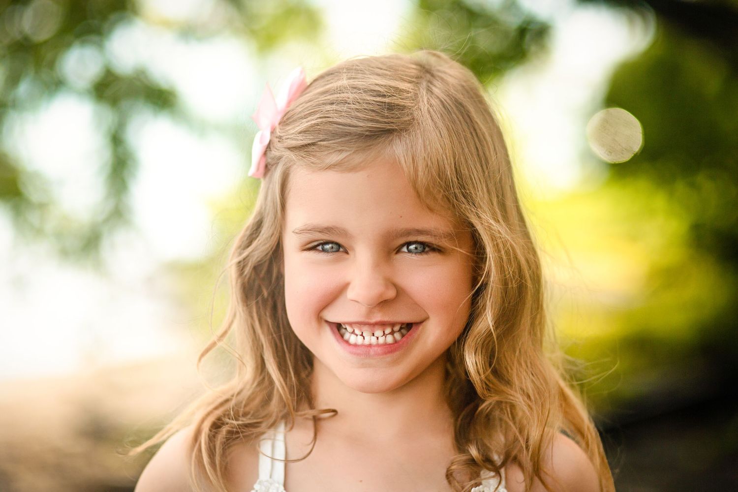 Smiling young girl with blonde hair and pink bow outdoors in soft natural light at the waterfront in Kingston, Ontario. Kingston Portraits.