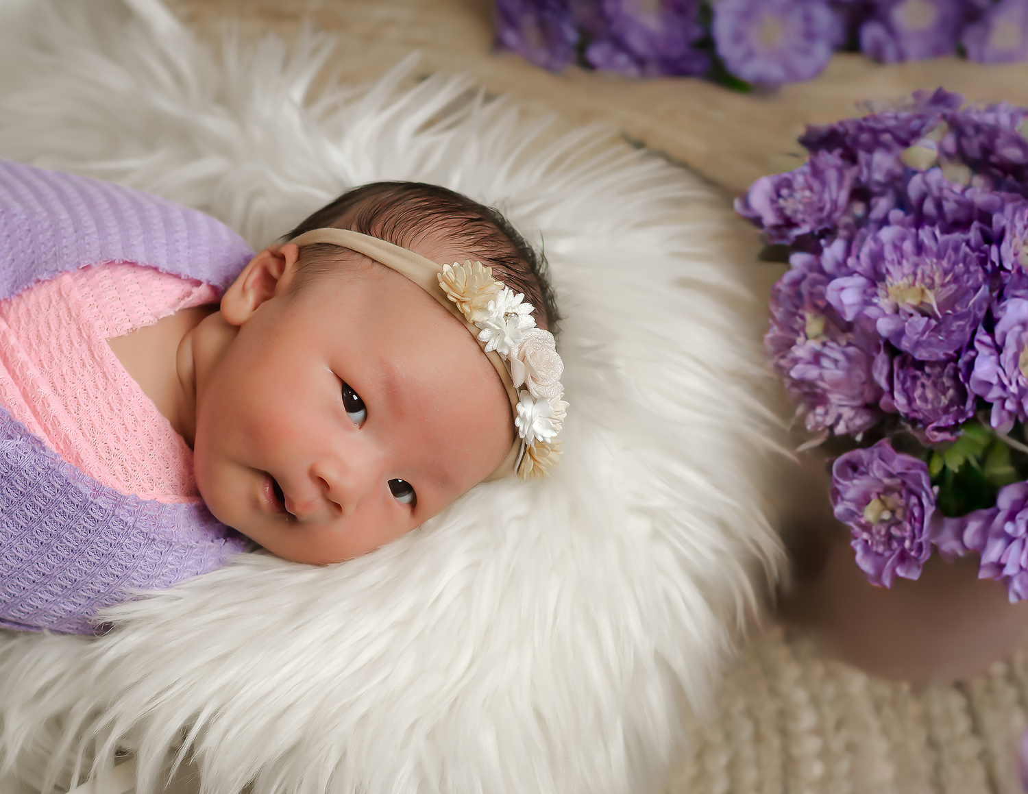 Newborn baby wrapped in pink and purple swaddle with floral headband on white fur surrounded by purple flowers, newborn photography studio.