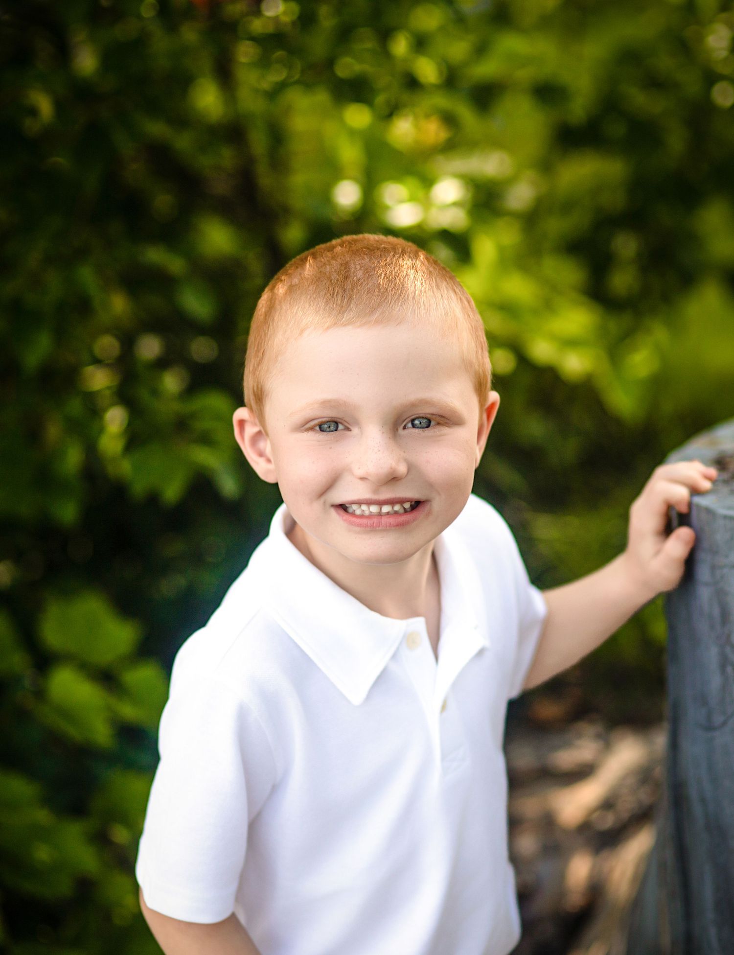 A smiling red-haired boy in a polo shirt poses outdoors with lush green foliage in the background at the waterfront in Kingston, Ontario.
