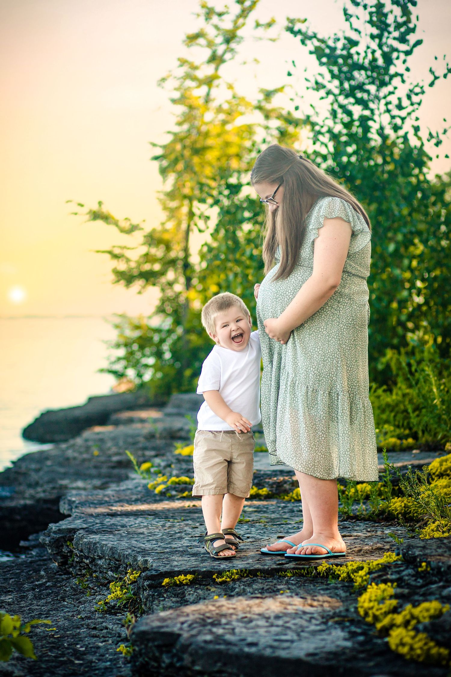 Pregnant mom in green dress smiling at her son on the rocky waterfront in Kingston, Ontario. Photos with Kingston Maternity photographer.