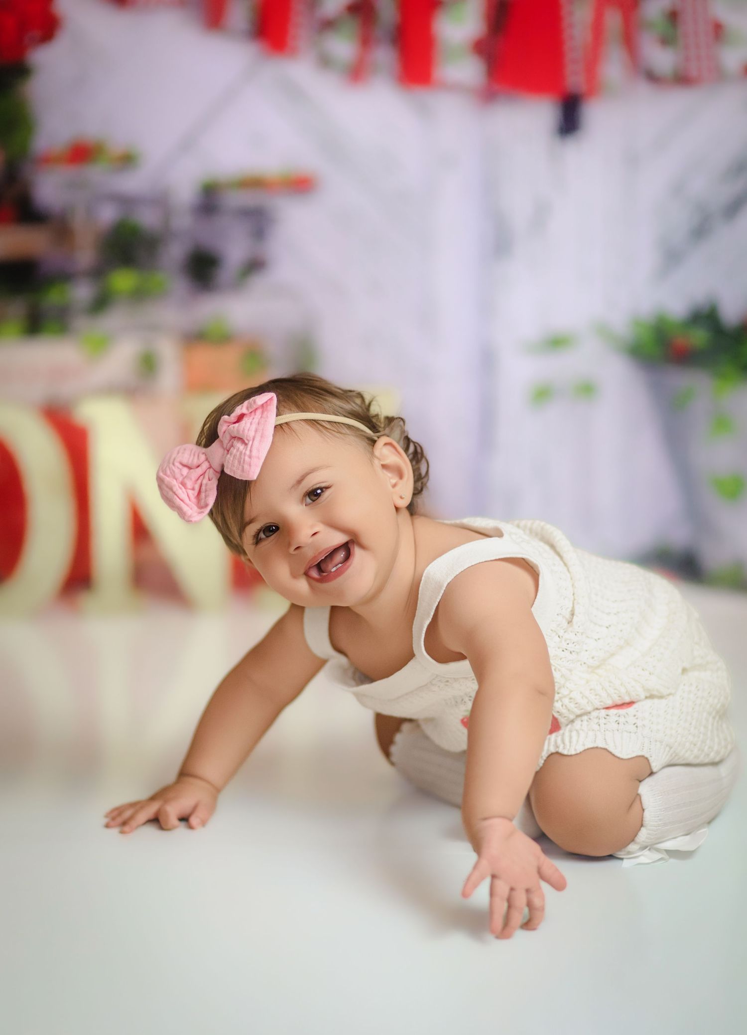 Smiling baby girl in white romper and pink bow headband crawling on floor during first birthday cake smash photoshoot in Kingston, Ontario.