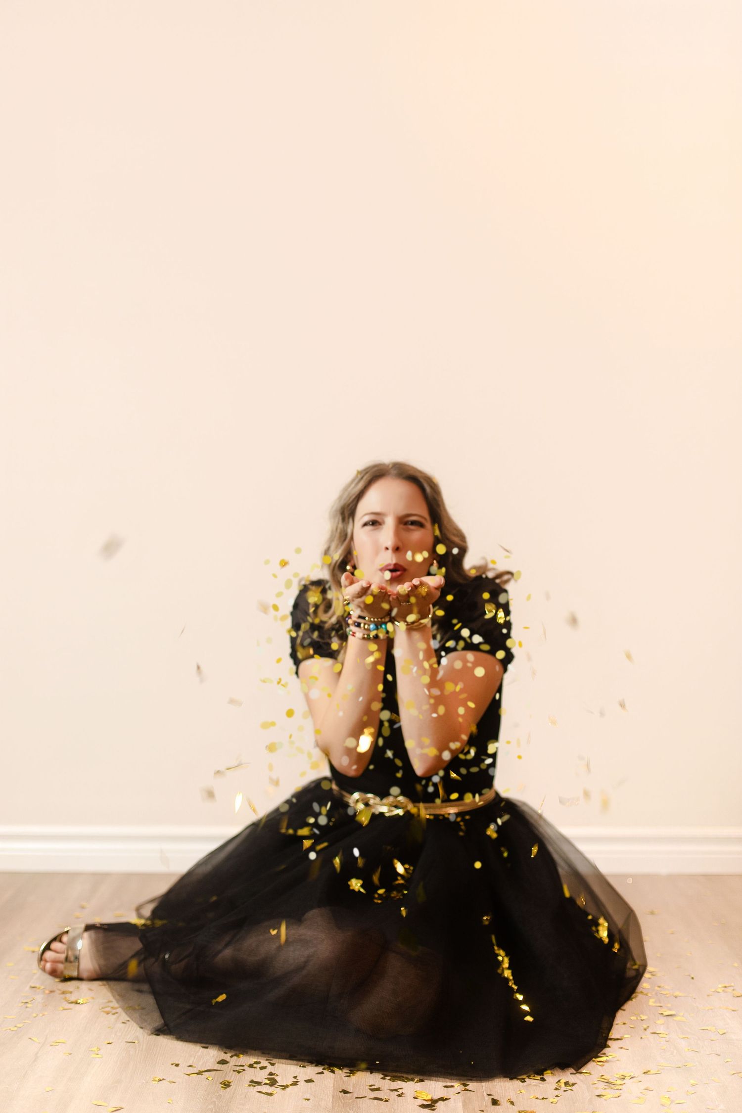 Woman in black tulle dress sitting and blowing gold confetti, celebrating in a bright and cheerful photography studio in Kingston, Ontario.