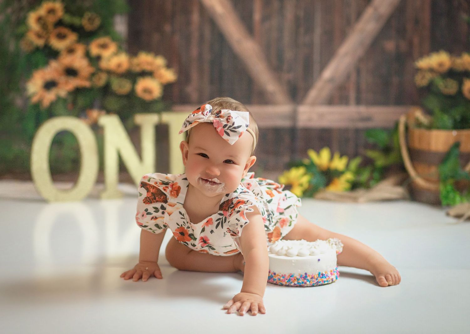 Baby girl in floral romper crawling near ONE sign and sunflowers during first birthday cake smash photo shoot in Kingston, Ontario.