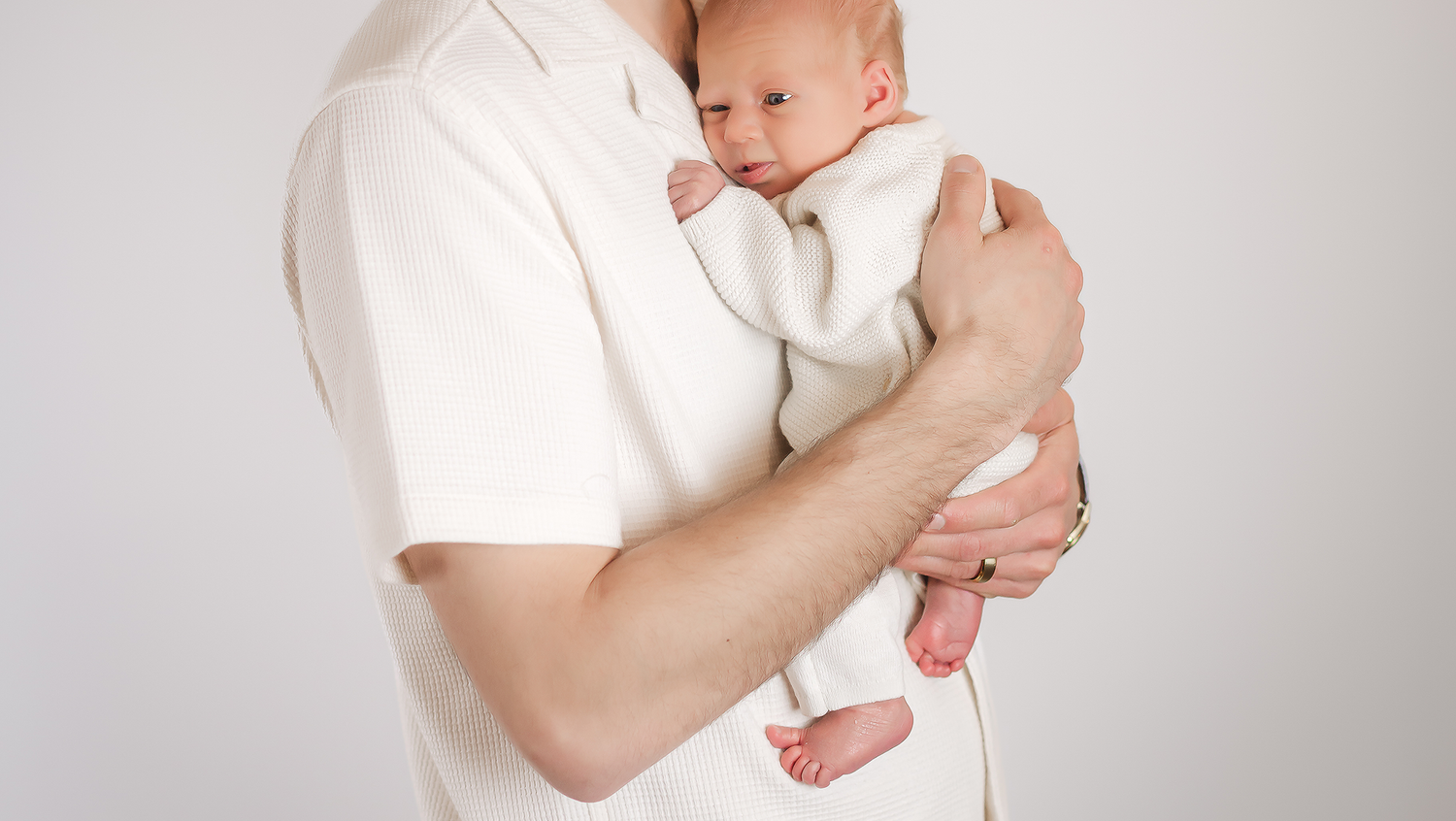 A father in a white shirt gently holds his newborn baby against a light background, newborn photo session in Kingston, Ontario.