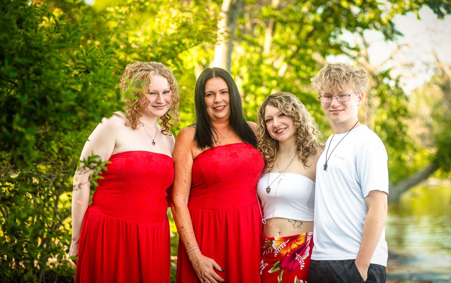 Family of four posing outdoors in red and white outfits, smiling among lush green trees near the waterfront in Kingston, Ontario.