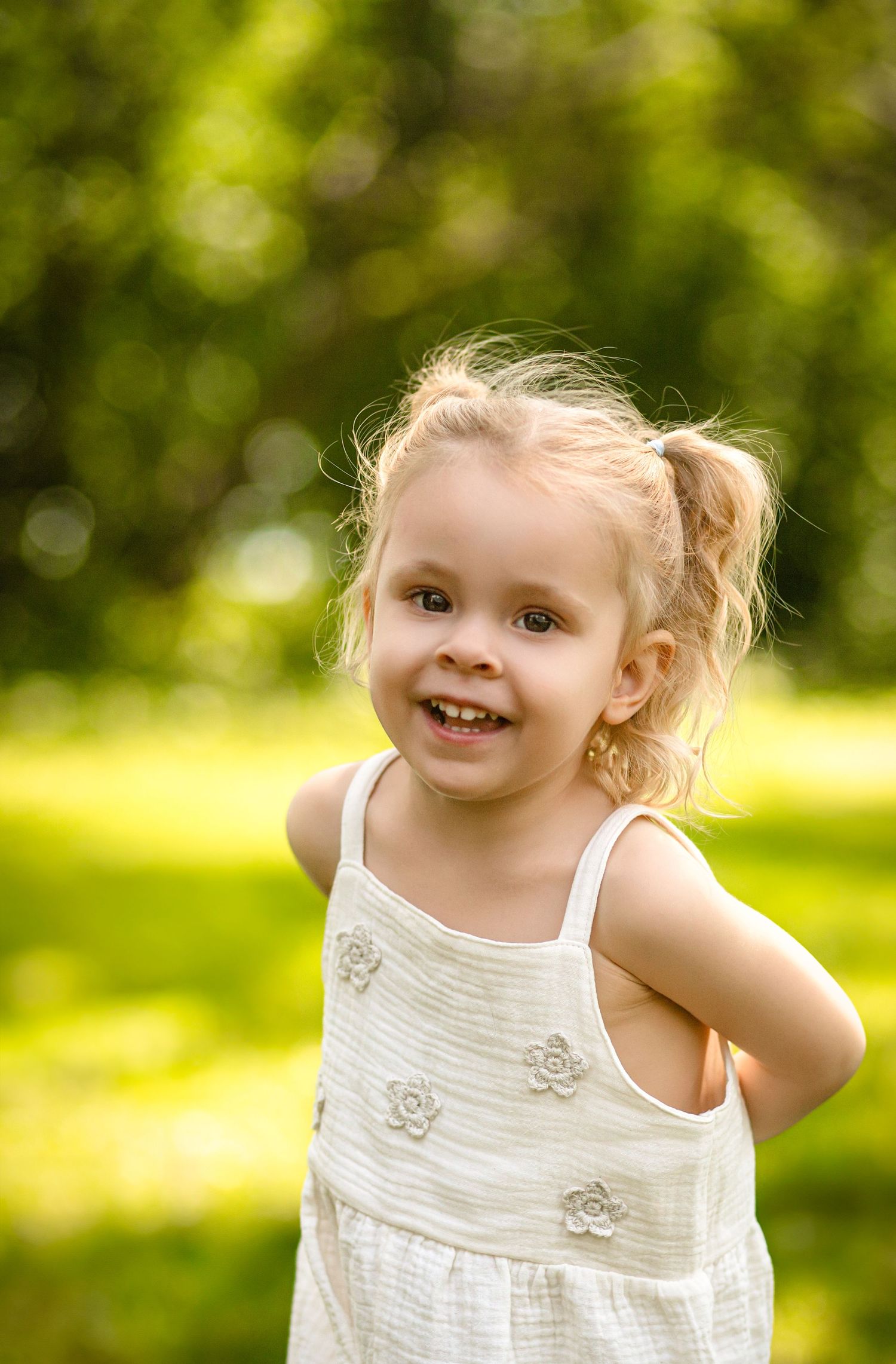 A cheerful toddler girl with pigtails wearing a white dress smiles outdoors in a sunny park in Kingston, Ontario, with family photographer.