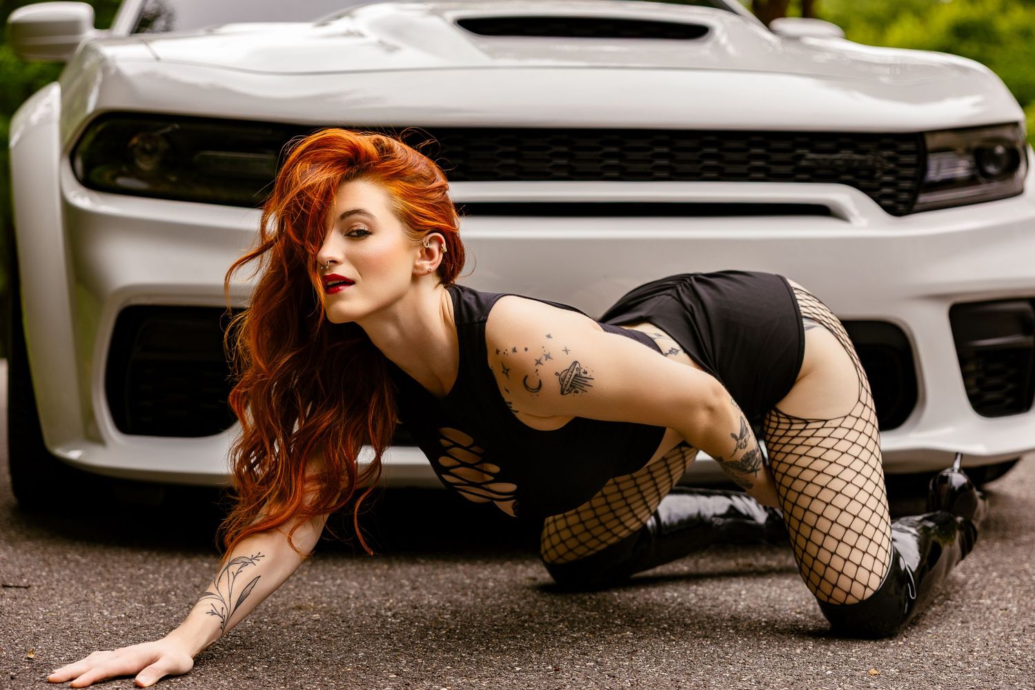 Redhead woman in black bodysuit and fishnet stockings posing in front of a white Dodge Charger on pavement.