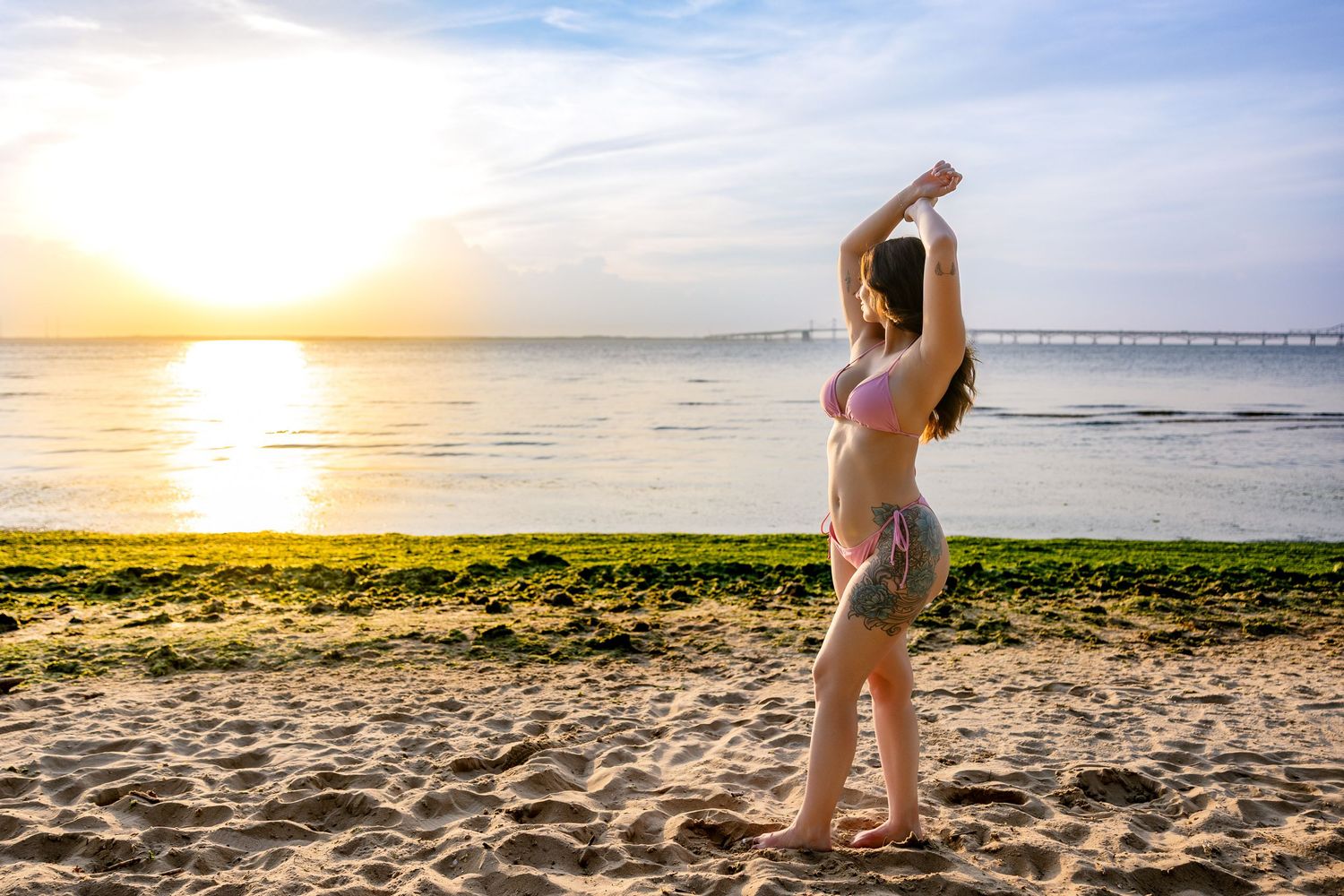 Woman in bikini stretching on sandy beach at golden sunset with ocean and pier in background.