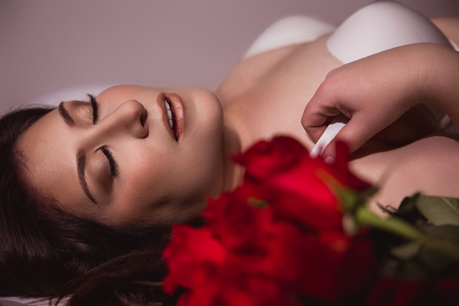 Woman lying down with red roses and a white flower petal held nearby, in a romantic boudoir setting.