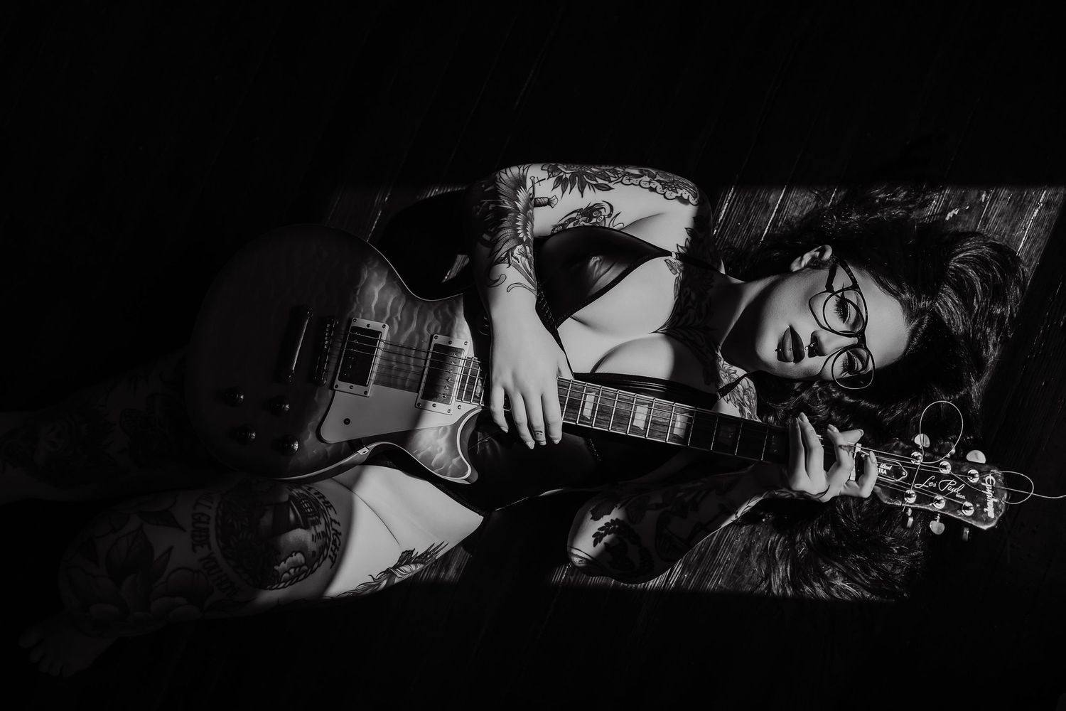 Black and white photo of a tattooed musician lying down playing an electric guitar while wearing glasses.
