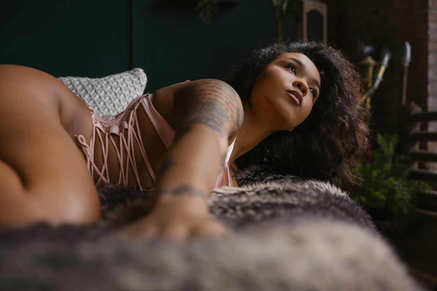 A tattooed woman with curly hair poses gracefully on a fur surface in a moody studio setting.