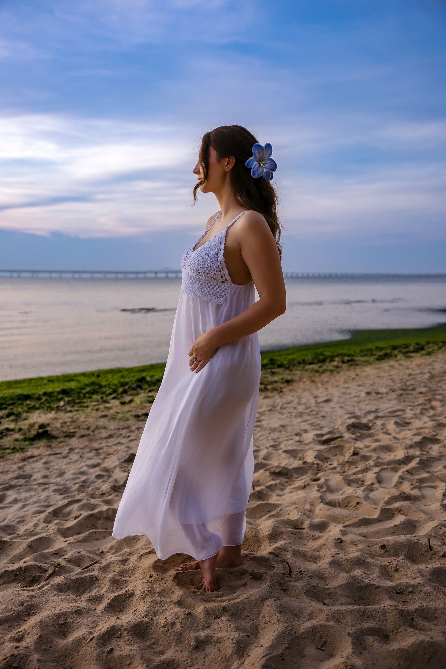 Woman in white maxi dress with blue flower hair accessory standing barefoot on sandy beach at dusk.