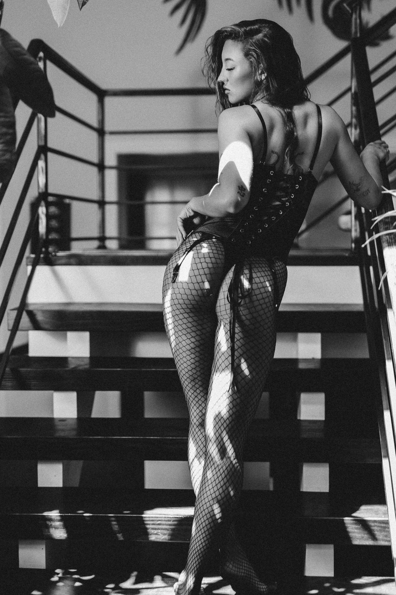 Black and white photo of a woman in fishnet tights and corset posing on outdoor stairs in sunlight.