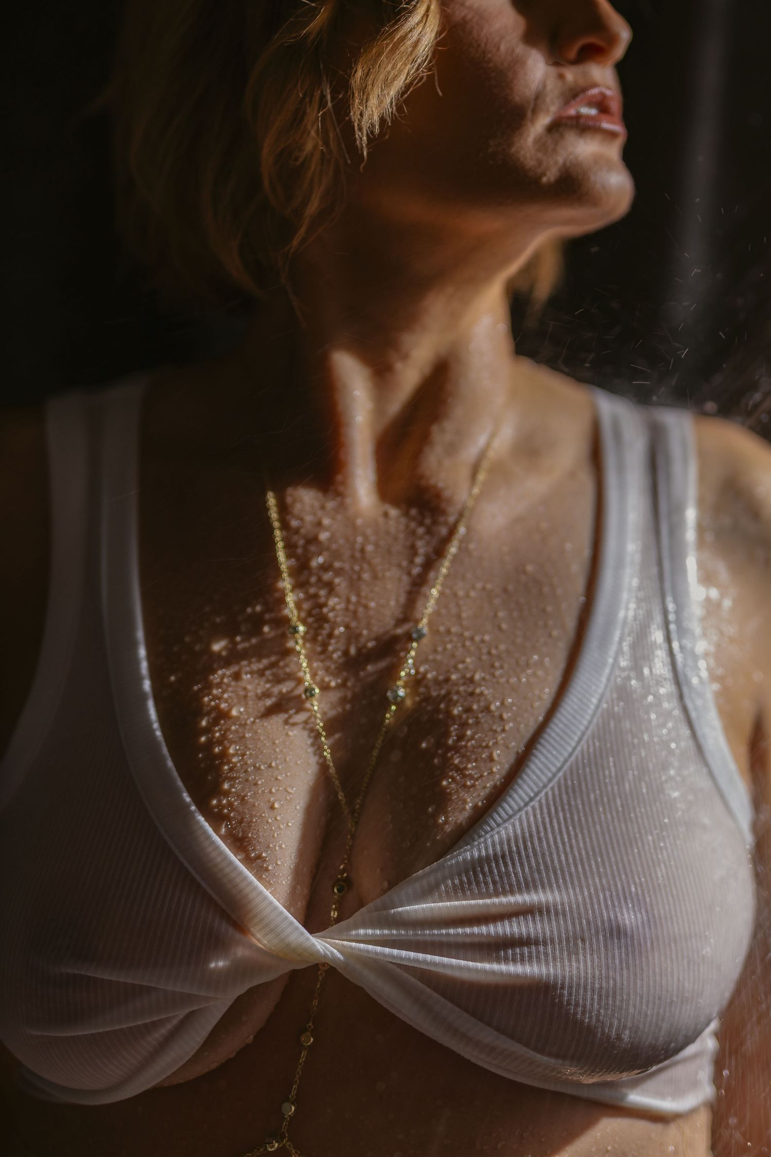 Artistic portrait showing water sprinkled across neck and chest area against dark background while wearing white tank top- boudoir wet set.