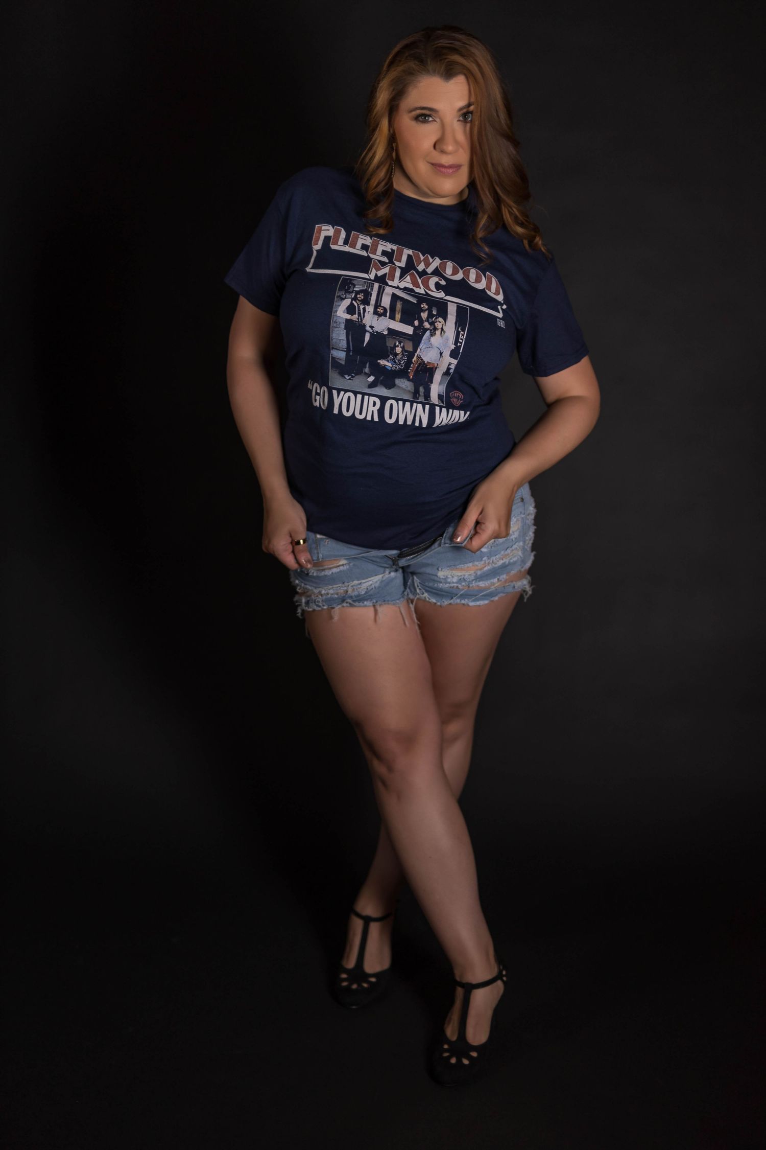 Woman in navy blue t-shirt and denim shorts poses against dark background in multiple casual poses.