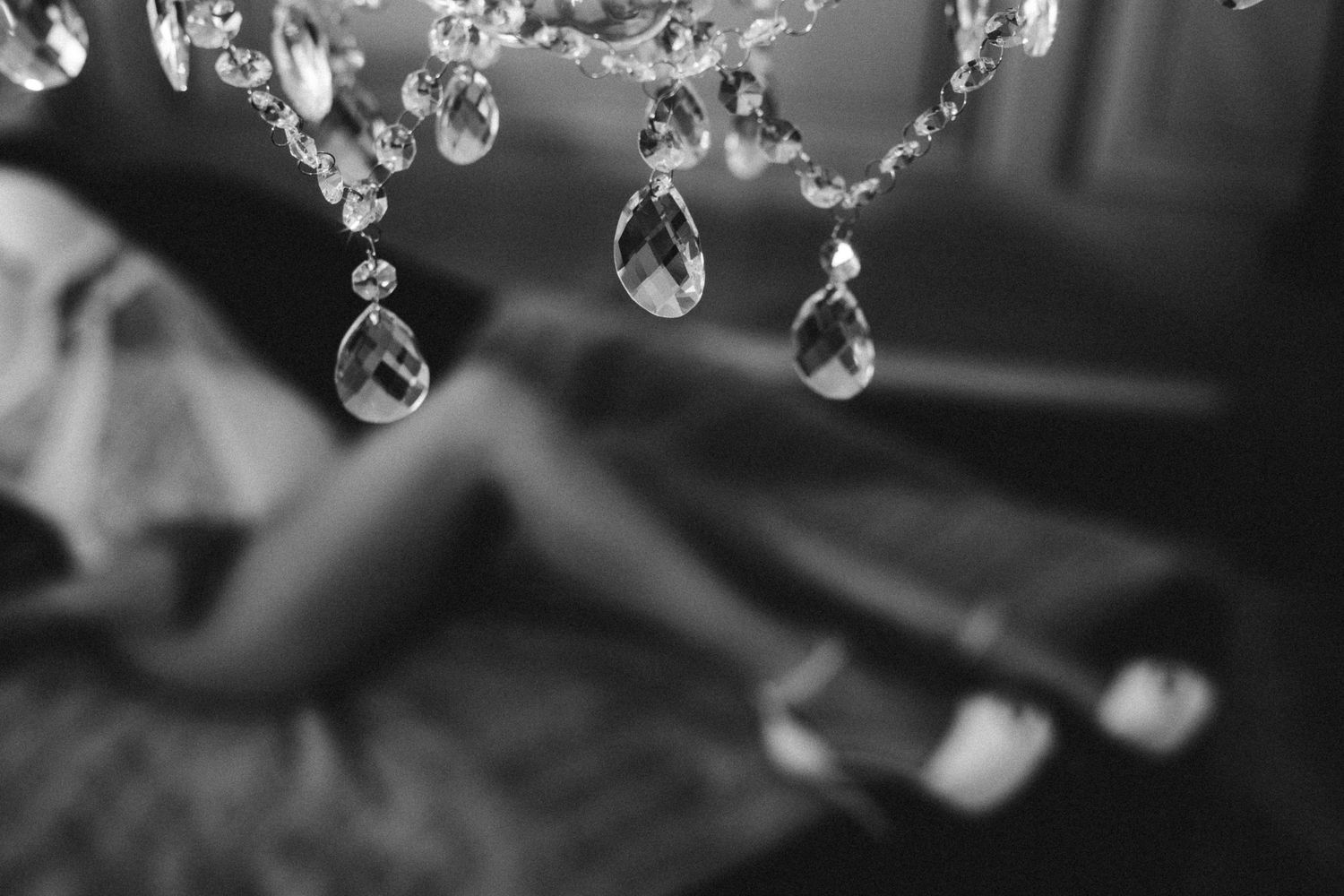 Black and white photo of a crystal chandelier with a blurred woman in heels resting on a couch in the background.