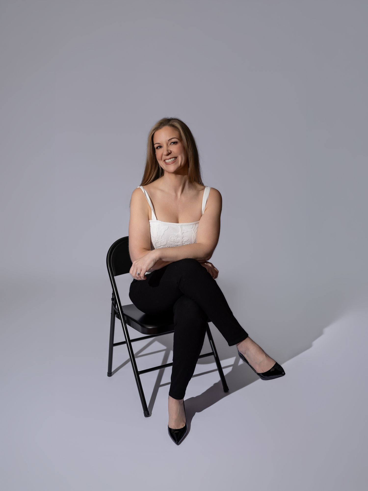 Woman in white top and black pants sitting on folding chair, smiling confidently against gray background.